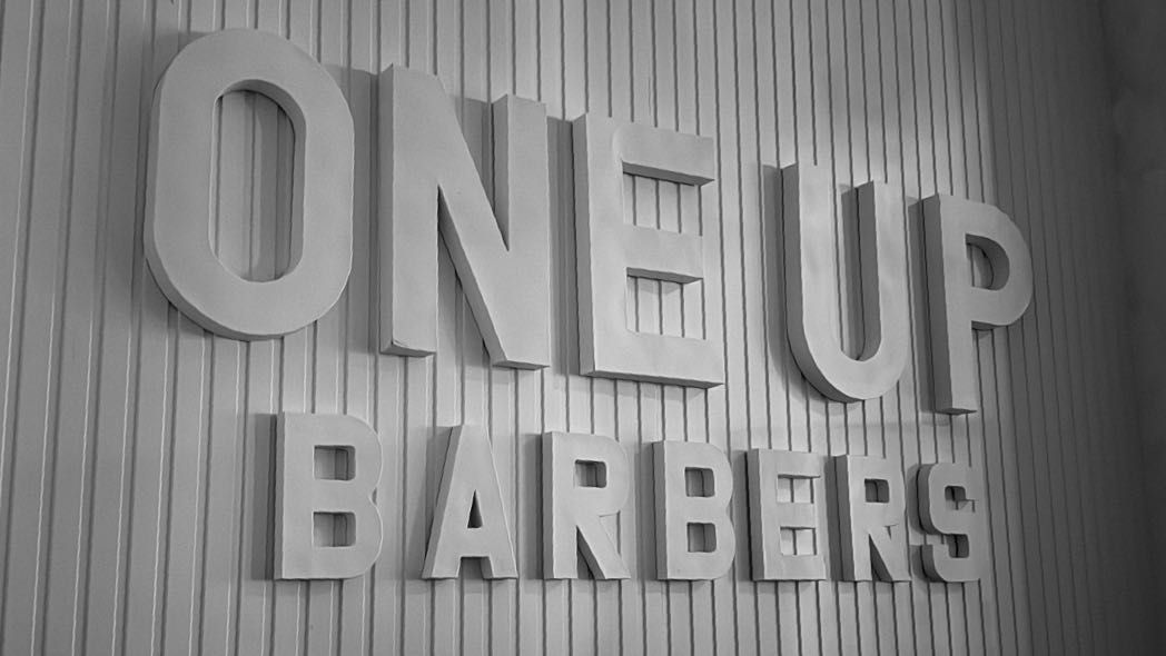 The dimensional ONE UP BARBERS lettering, cut in white and mounted on a white fluted panel.