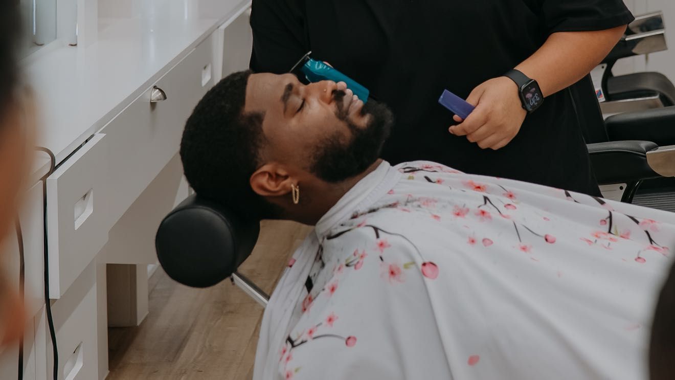 A client reclined in the barber chair mid cut, wearing a floral cape, beside the white station cabinetry.